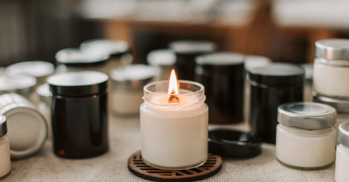 Gros plan d'une bougie blanche artisanale avec une flamme enflammée, entourée de pots de bougies sur une table.