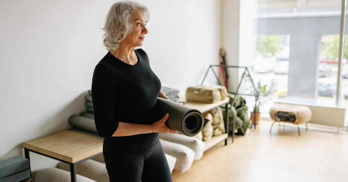 Femme âgée en tenue noire tenant un tapis de yoga dans un espace lumineux et minimaliste.